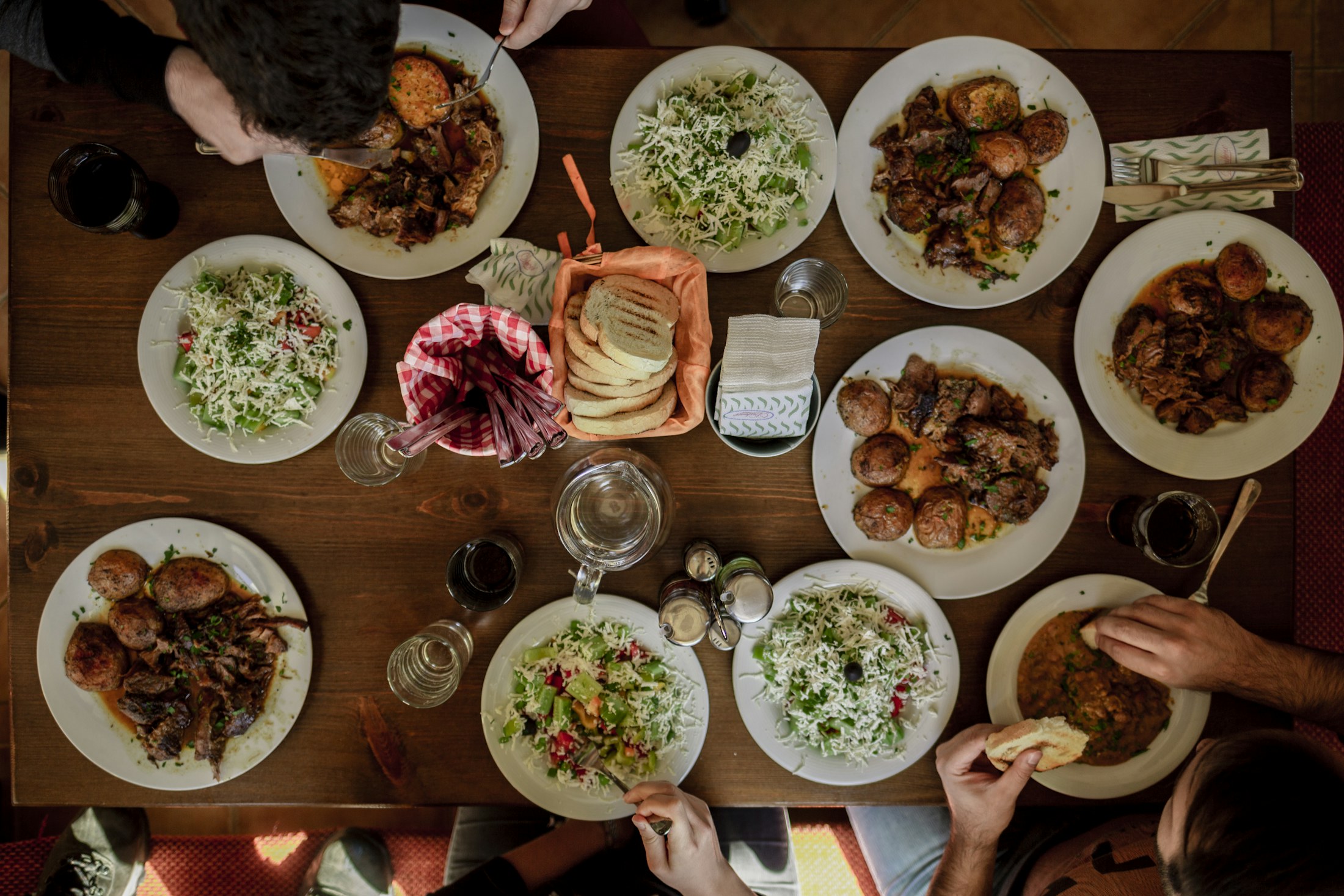 An overhead view of a long wooden table crowded with plates of warm home-cooked food, with several people's hands reaching in to share the meal